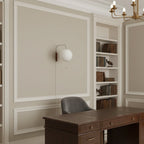 Small round Clérachic wall light on a panelled wall in a study, with built-in shelves, a dark wood desk and a grey chair below.