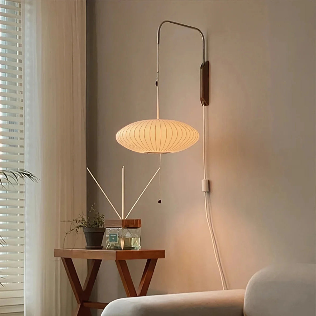 A wide, flat saucer-shaped glowing shade hangs from a tall Clérachic wall arm beside a folding wooden side table and a curtained window.