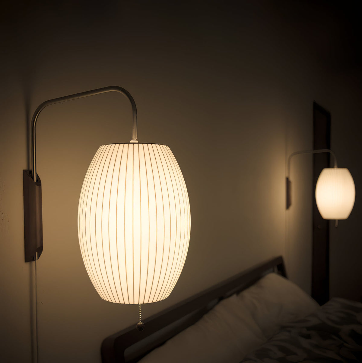 Close view of a glowing oval ribbed shade on this Clérachic wall light in a dim bedroom, with a second lit shade behind it.