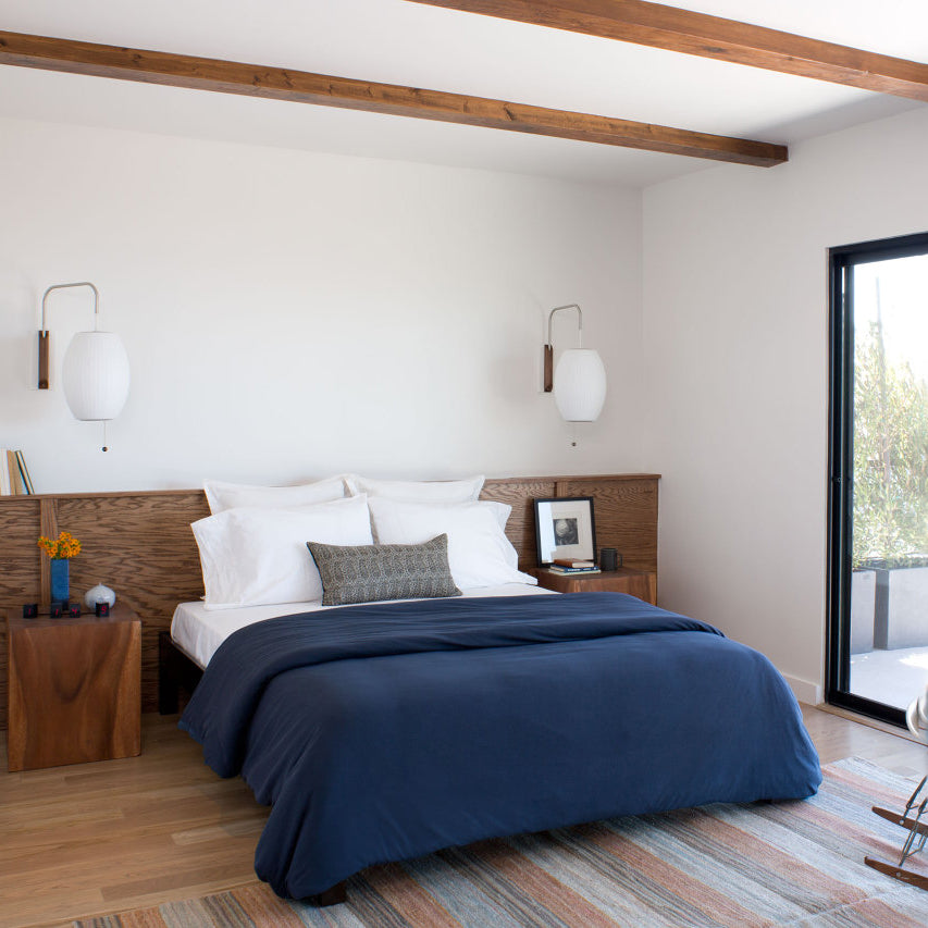 Pair of Clérachic wall lights with oval white shades above a bed with navy bedding, a wood headboard and a balcony door beside it.