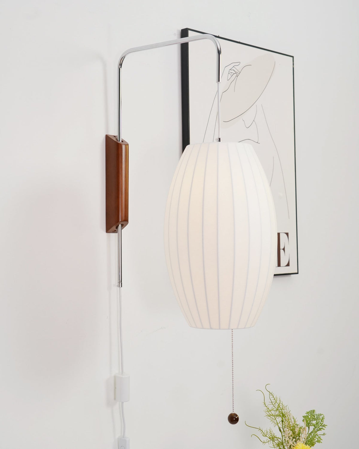 Oval white shade of this Clérachic wall light hangs in front of a framed line drawing, with a small vase of yellow flowers at the bottom.