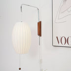 Close view of the oval shade, steel arm and walnut bracket of this Clérachic wall light, with the edge of a framed print behind.