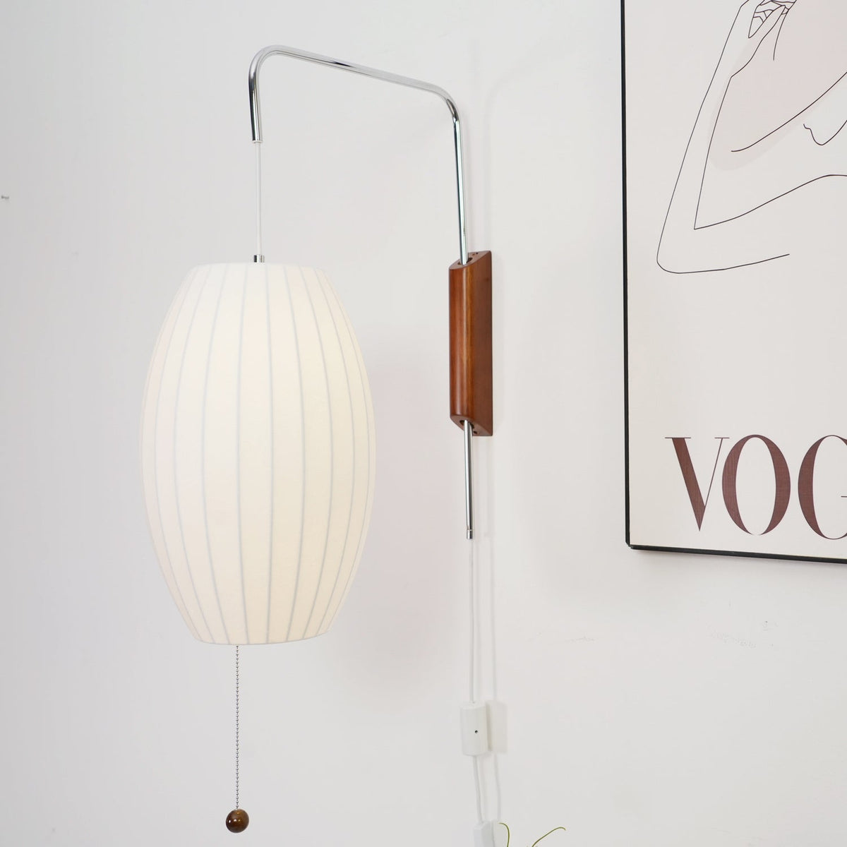 Close view of the oval shade, steel arm and walnut bracket of this Clérachic wall light, with the edge of a framed print behind.