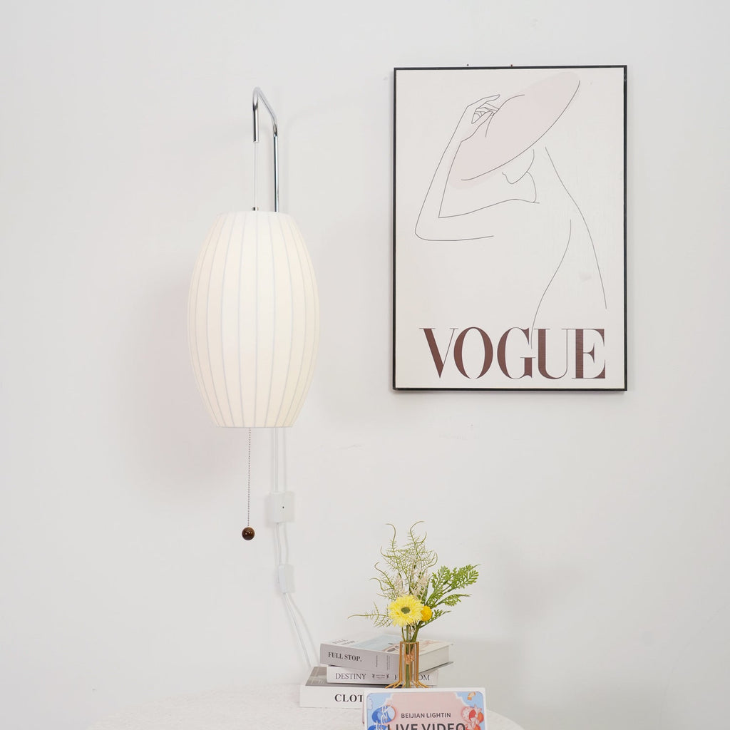 A Clérachic wall light with a slim oval shade hangs beside a framed line drawing, with a small vase of flowers and a stack of books below.