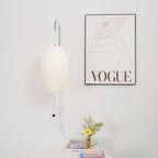 A Clérachic wall light with a slim oval shade hangs beside a framed line drawing, with a small vase of flowers and a stack of books below.