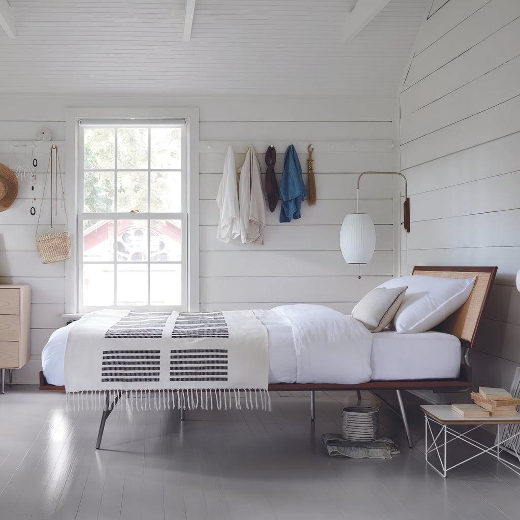 Oval white shade of a Clérachic wall light hangs beside a window above a low bed in a white-boarded room, with coats on pegs.