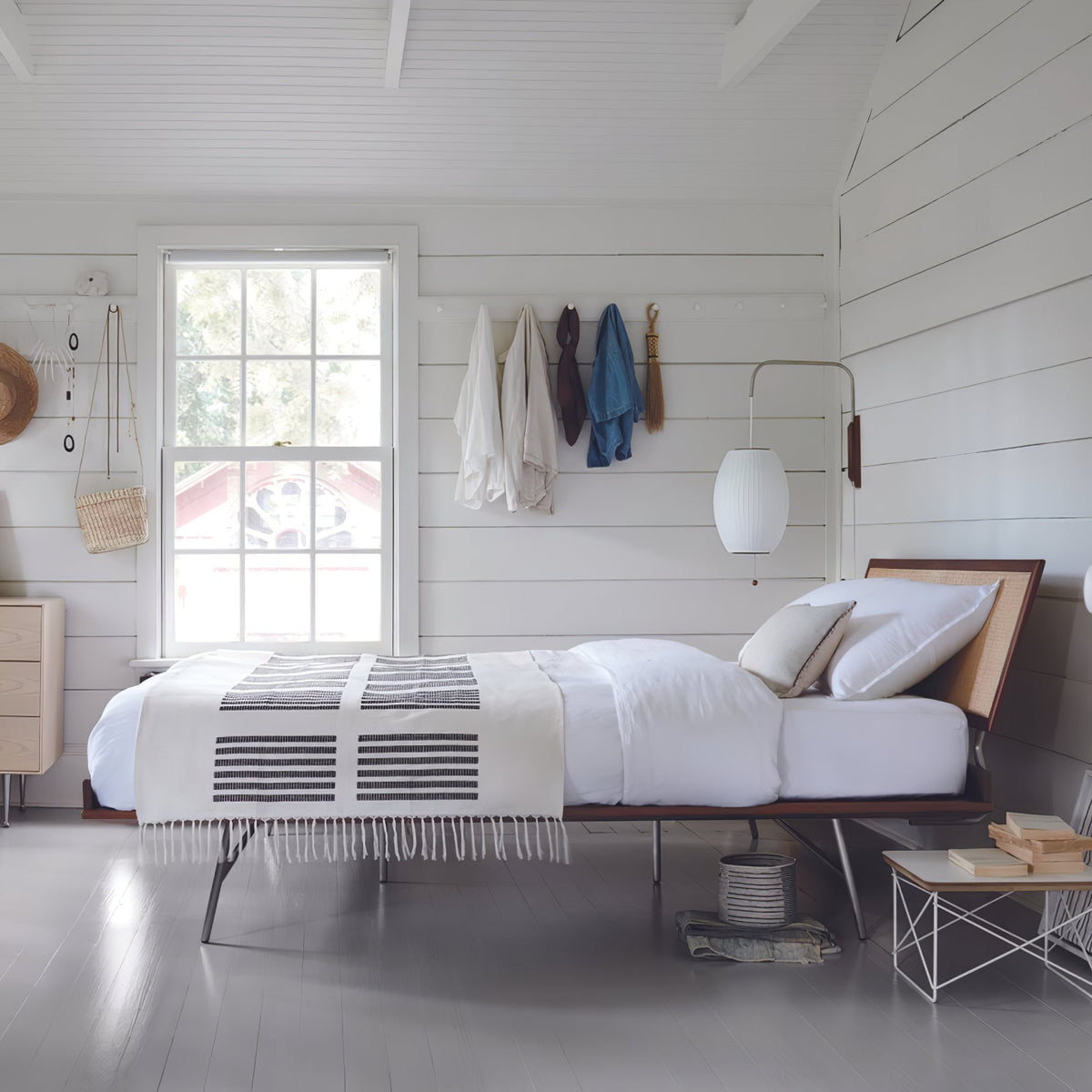 Oval white shade of a Clérachic wall light hangs beside a window above a low bed in a white-boarded room, with coats on pegs.