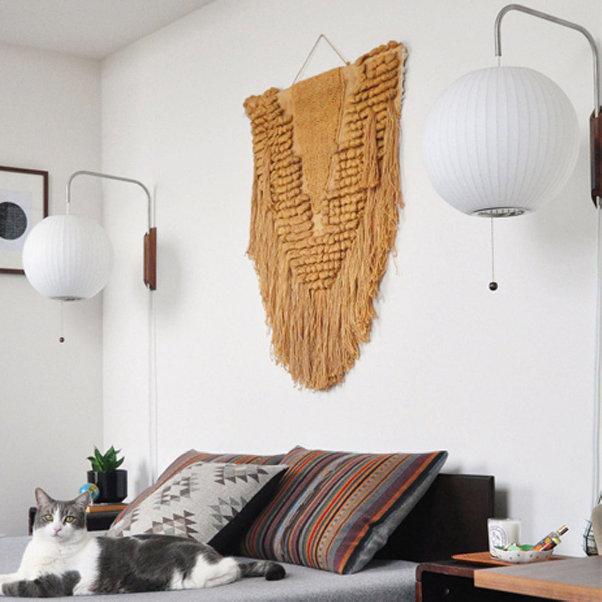 Two round Clérachic wall lights of different sizes flank an ochre woven wall hanging above a daybed where a cat rests.