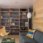 An oval Clérachic wall light on a blonde wood wall, set beside built-in bookshelves with a grey sofa and a yellow butterfly chair.
