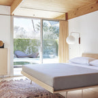 A round Clérachic wall light on a short arm beside a sliding glass door, above a low platform bed with grey bedding.