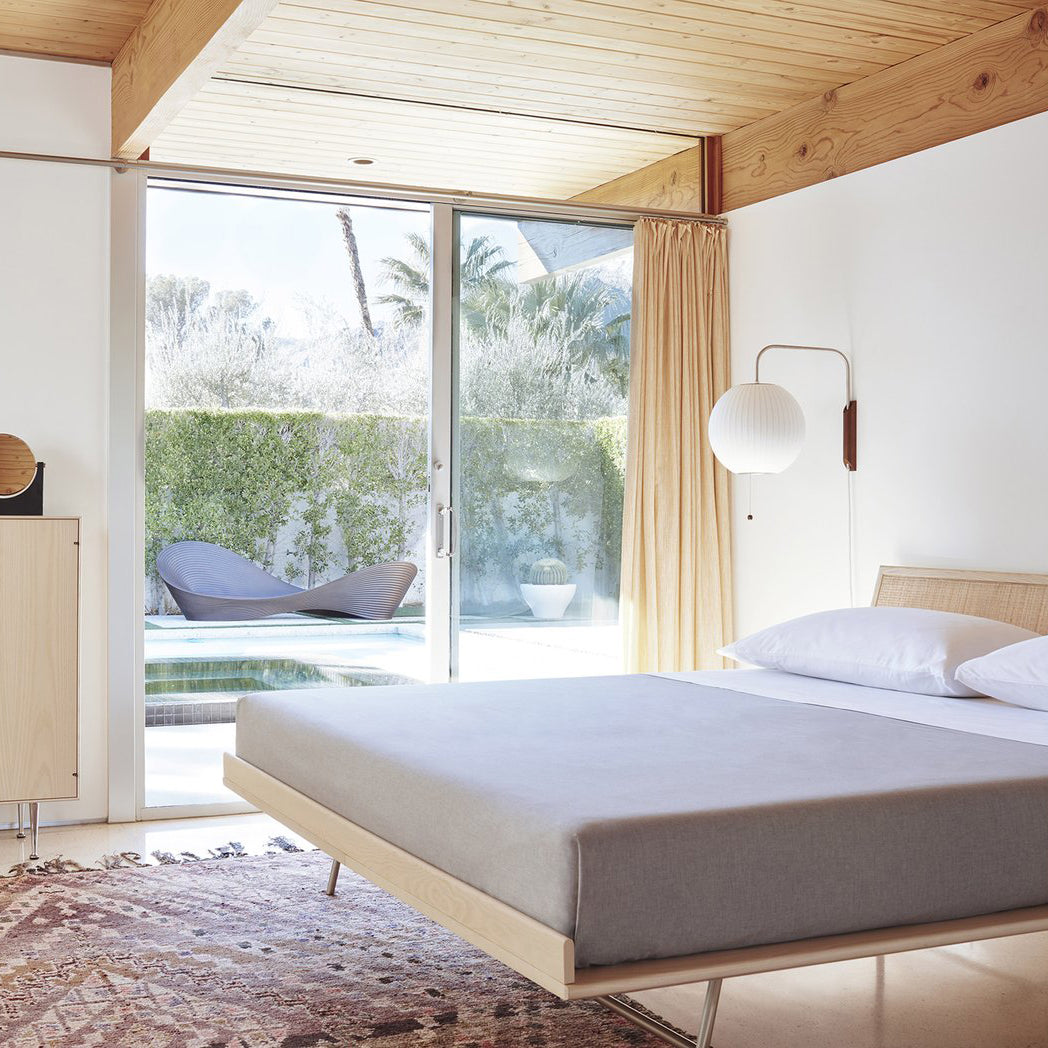 A round Clérachic wall light on a short arm beside a sliding glass door, above a low platform bed with grey bedding.