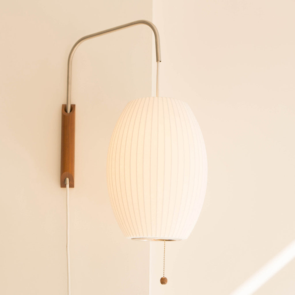 Warm light fills the oval shade of this Clérachic wall light, with a walnut bracket and pull cord against a peach-toned wall.