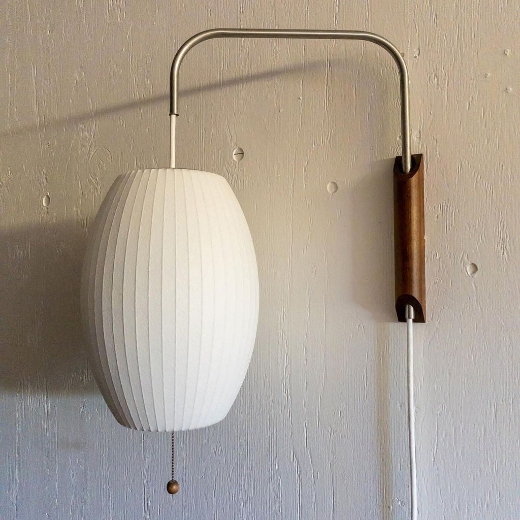 Close view of the ribbed oval shade on a Clérachic wall light, with its steel arm, walnut bracket and wooden pull bead on a textured grey wall.