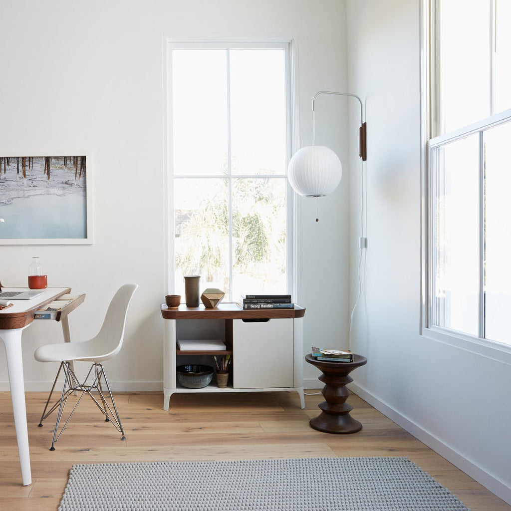 A round Clérachic wall light sits high on the wall of a bright room, near tall windows, a desk and a small cabinet.