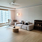 Small Clérachic wall light glowing beside a window in a spacious living room, with a pale sofa, a round wood table and a ceiling fan.