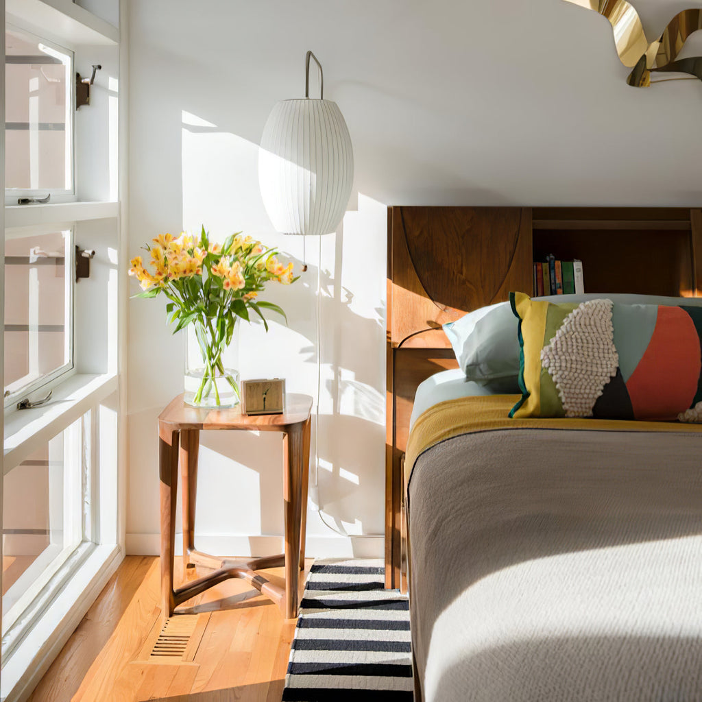 A tall oval Clérachic wall light hangs beside a window and a bed with colourful cushions, above a walnut side table with yellow flowers.