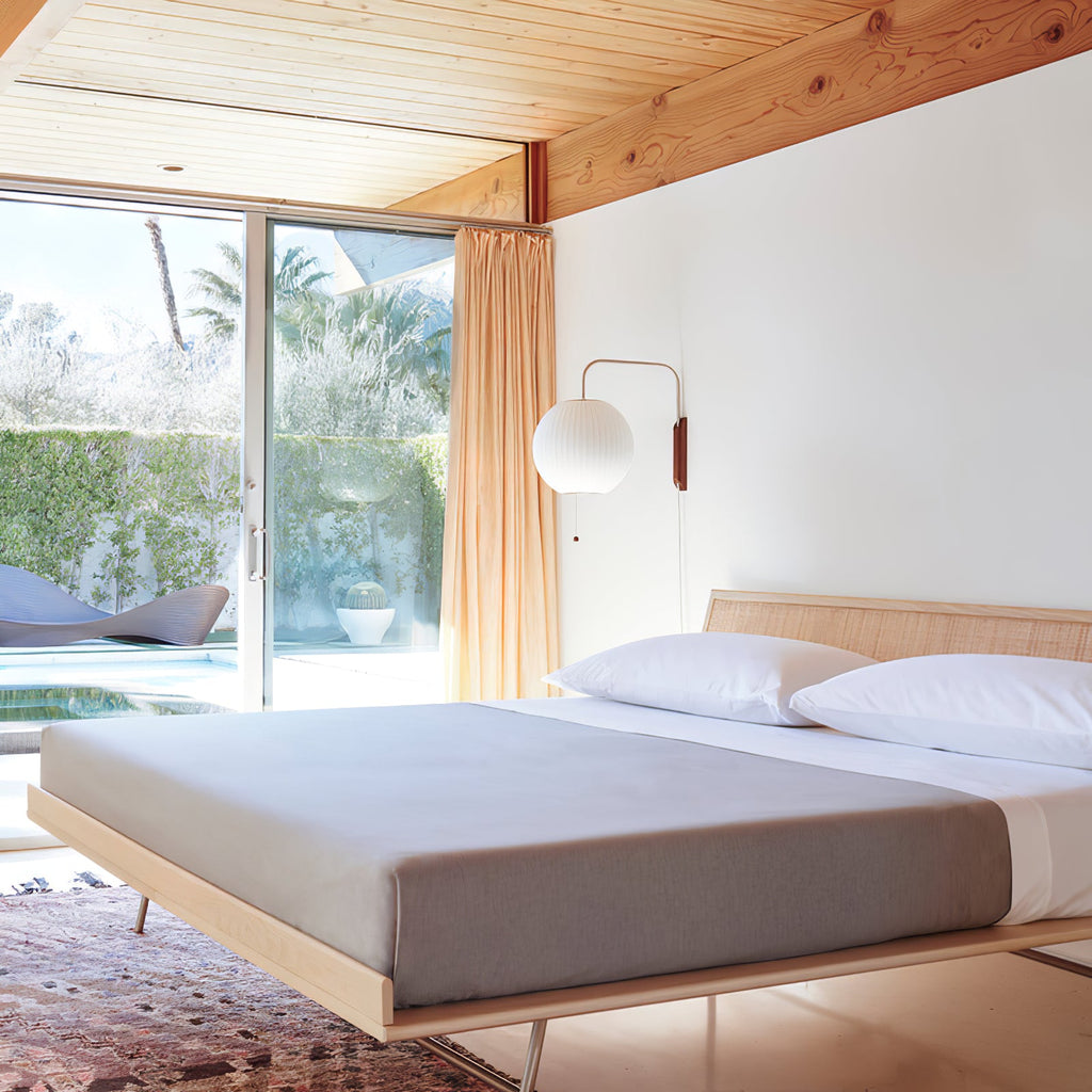 A round Clérachic wall light hangs beside a sliding glass door above a platform bed with grey bedding, under a wooden ceiling.