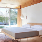 A round Clérachic wall light hangs beside a sliding glass door above a platform bed with grey bedding, under a wooden ceiling.