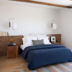 Two oval Clérachic wall lights on either side of a wood-panelled headboard, above a bed with navy bedding and a patterned cushion.
