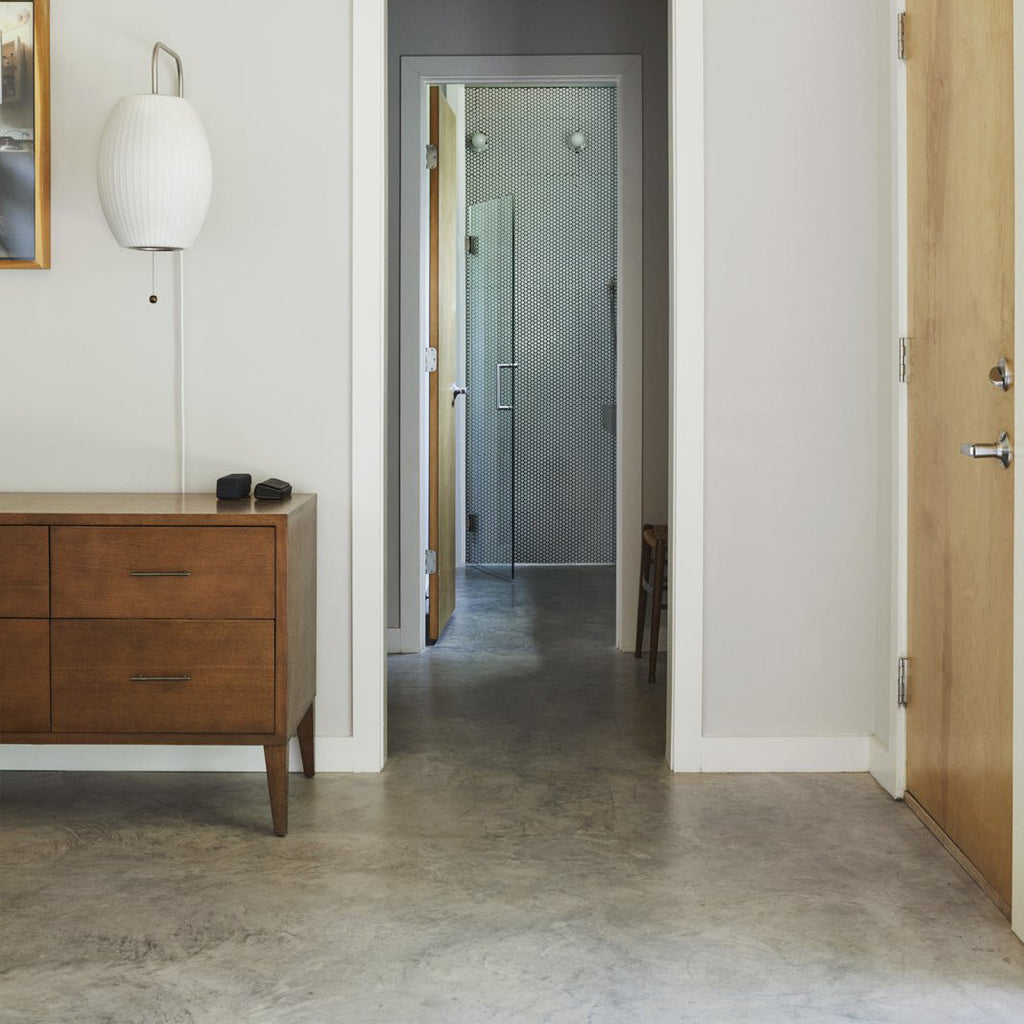 A round white Clérachic wall light at the top left of a hallway scene, above a walnut chest of drawers and a concrete floor.
