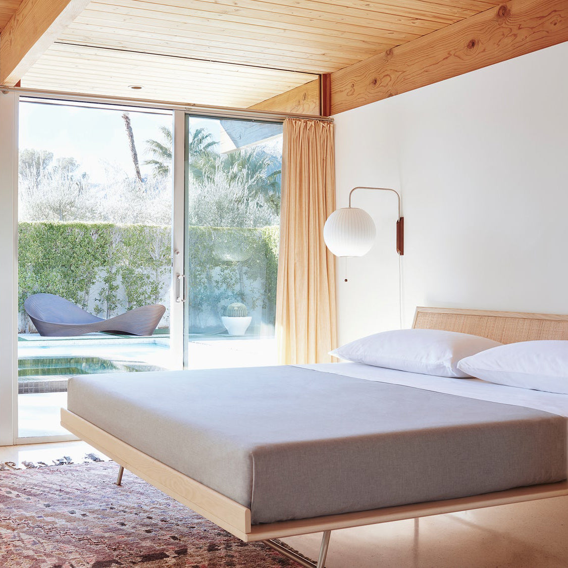 Round white Clérachic wall light beside a sliding door and orange curtain, above a low platform bed and a patterned rug.
