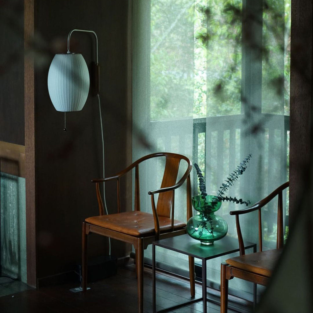 Pale oval shade of a Clérachic wall light in a dark, leafy room, beside two wooden chairs and a glass vase on a small table.