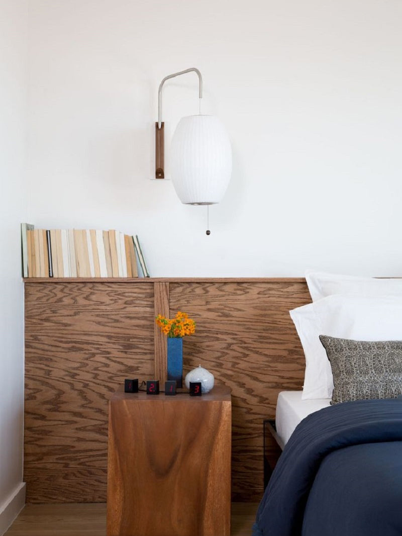 Oval Clérachic wall light above a wooden bedside cube with a vase of yellow flowers, next to a navy bed throw and a plywood headboard.