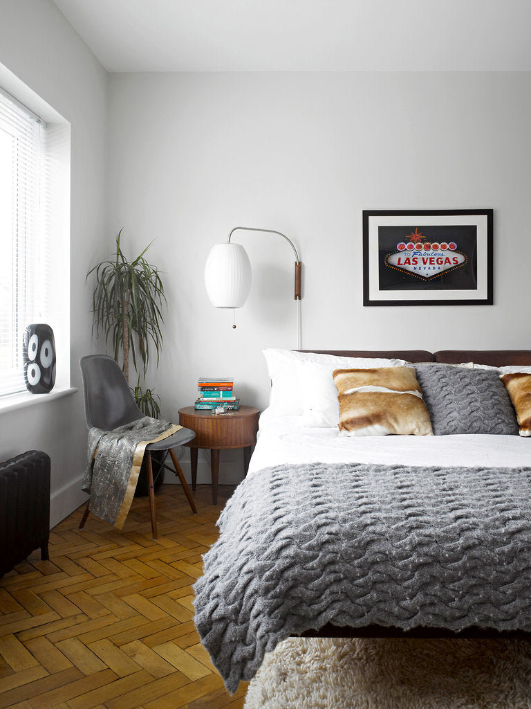 A Clérachic wall light with a small round shade hangs above a bed with a grey knitted throw, beside a window and a framed print.