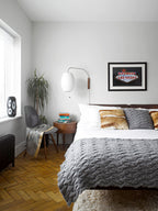 A Clérachic wall light with a small round shade hangs above a bed with a grey knitted throw, beside a window and a framed print.