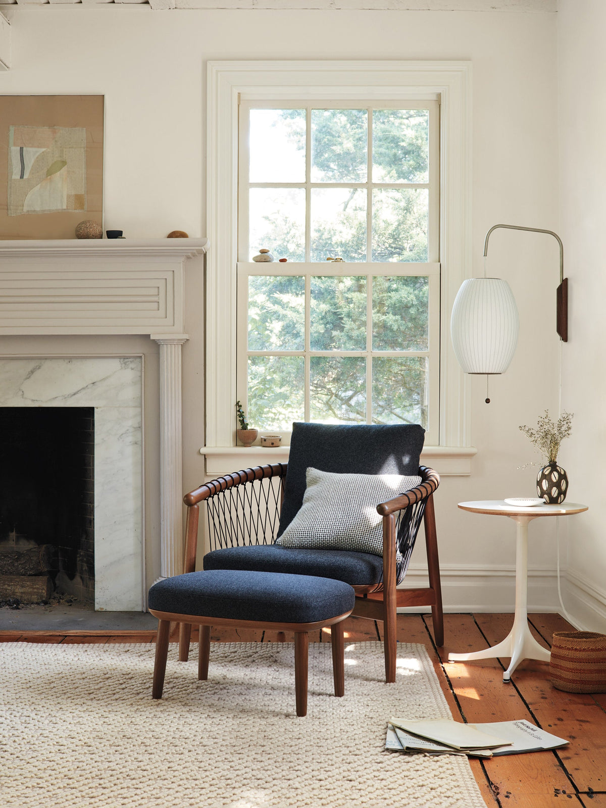 Oval Clérachic wall light beside a sash window and fireplace, with a dark armchair, footstool and a pale rug in the foreground.