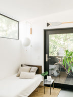 Small round Clérachic wall light by a glass door in a bright room, above a single bed with cushions, with a wooden paddle on the wall.