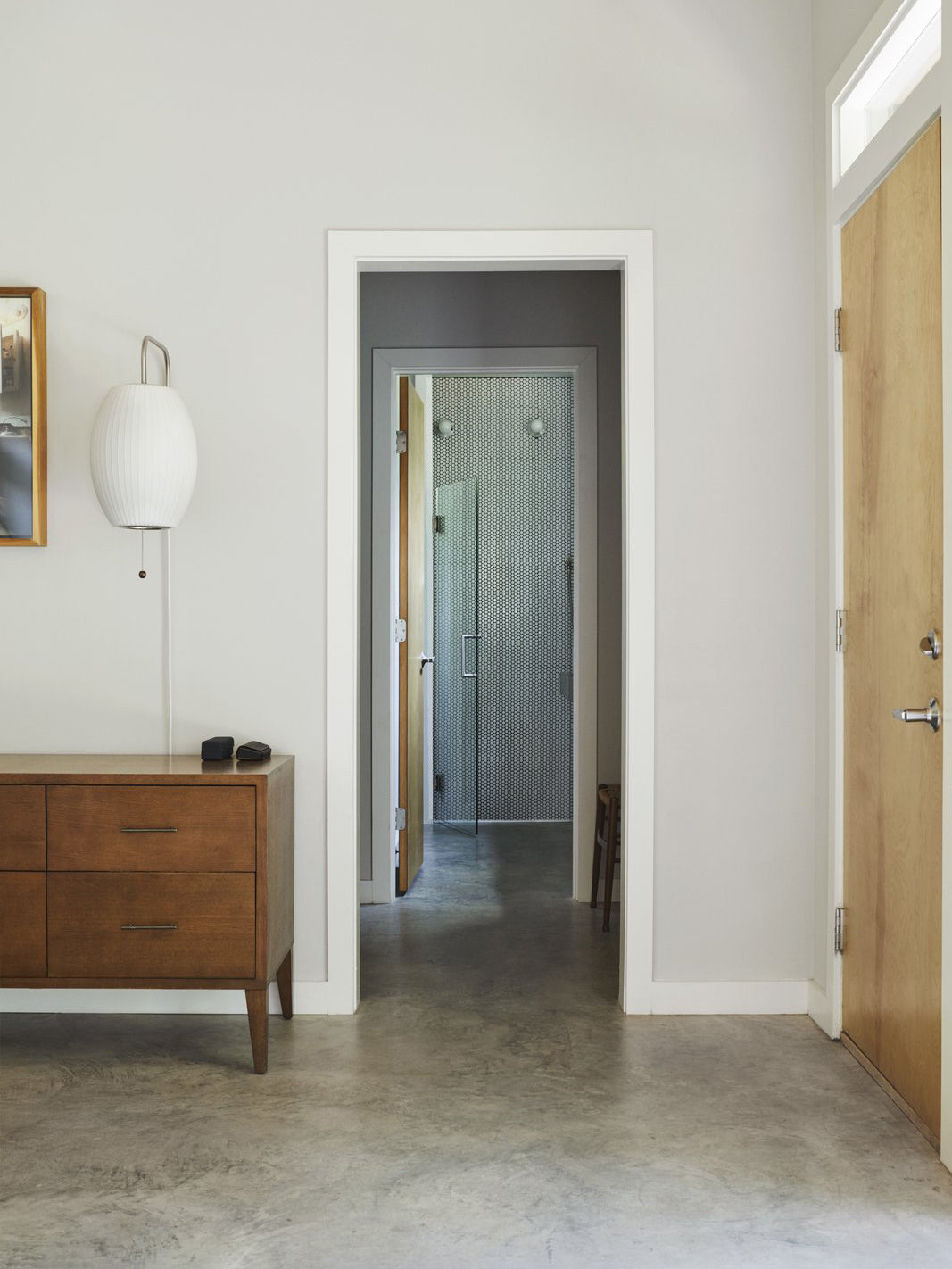 Round white Clérachic wall light high on a pale hallway wall, above a walnut chest of drawers, with a door opening to a corridor.