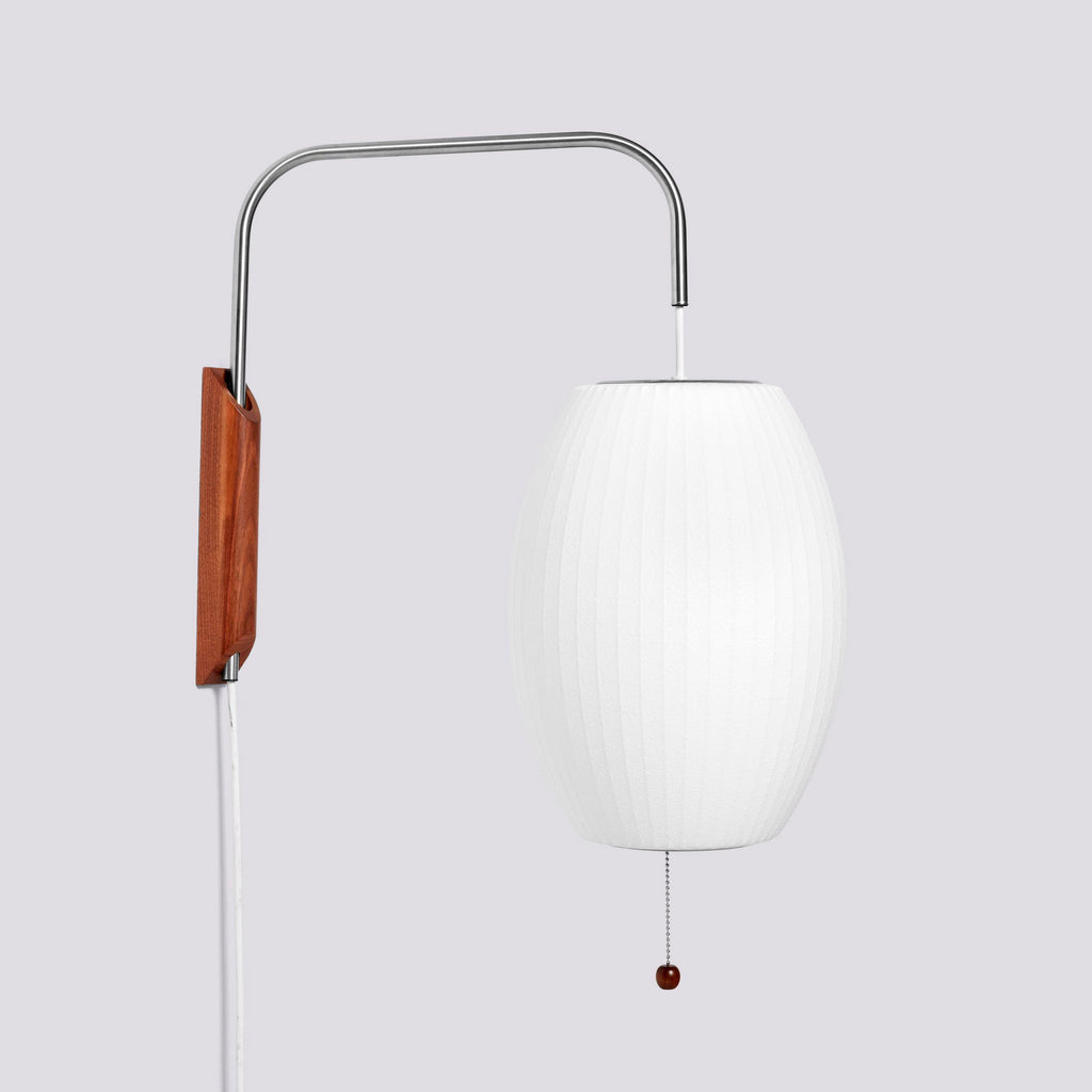 Oval white shade of this Clérachic wall light hangs from a curved steel arm beside its walnut bracket, on a light grey backdrop.