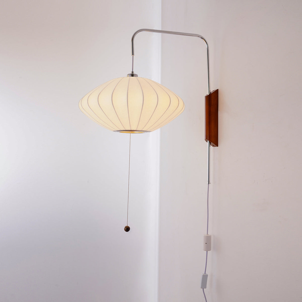 Lit saucer-shaped shade glowing warm on a Clérachic wall light, with its pull cord, small in-line switch and white cable on a pale wall.