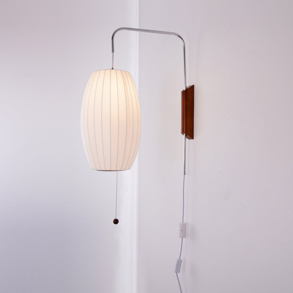 Oval shade of a Clérachic wall light lit warm, with a walnut bracket, pull cord and in-line switch against a pale wall.