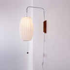 Oval shade of a Clérachic wall light lit warm, with a walnut bracket, pull cord and in-line switch against a pale wall.