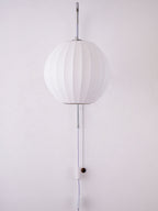 Round white shade of a Clérachic wall light, unlit, with its cable and wooden bead running down a pale wall.