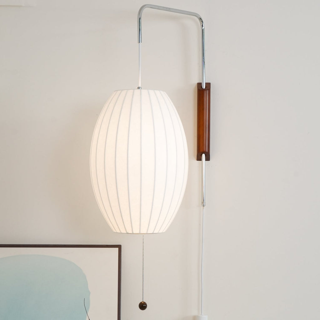 A larger lit oval shade hangs from this Clérachic wall light, with a walnut bracket and bead pull above a framed artwork.