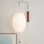 A larger lit oval shade hangs from this Clérachic wall light, with a walnut bracket and bead pull above a framed artwork.