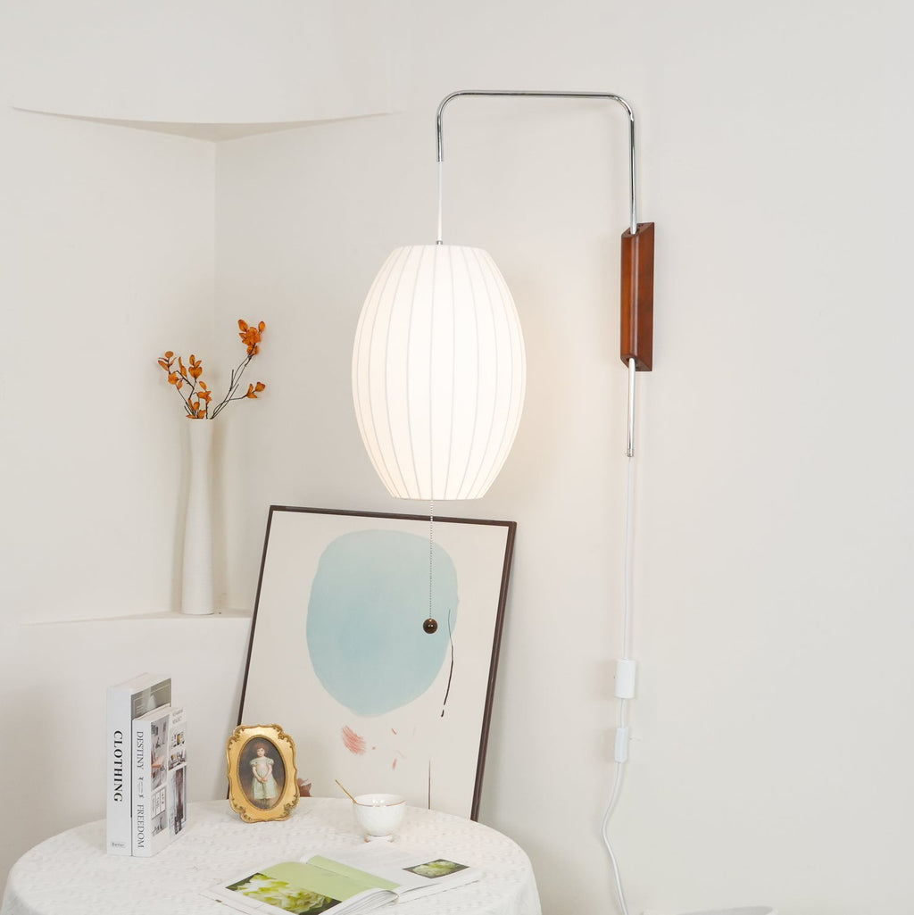 Glowing oval shade on a Clérachic wall light in a cream corner, above a round table with books, a gold frame and a vase of orange sprigs.