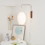Glowing oval shade on a Clérachic wall light in a cream corner, above a round table with books, a gold frame and a vase of orange sprigs.