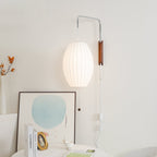 A Clérachic wall light with a lit oval shade in a cream corner, with a framed blue artwork and a vase of autumn sprigs below.