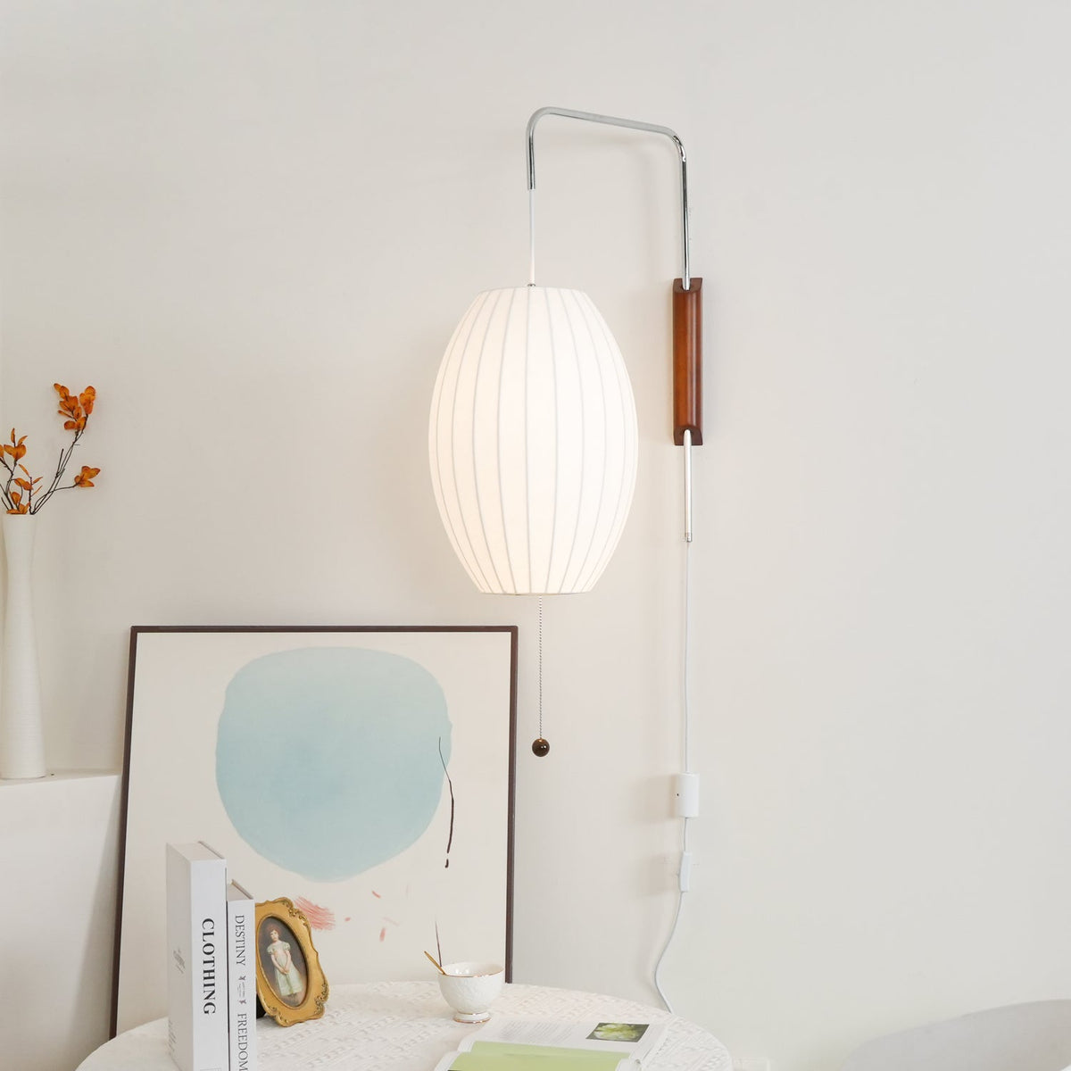 A Clérachic wall light with a lit oval shade in a cream corner, with a framed blue artwork and a vase of autumn sprigs below.
