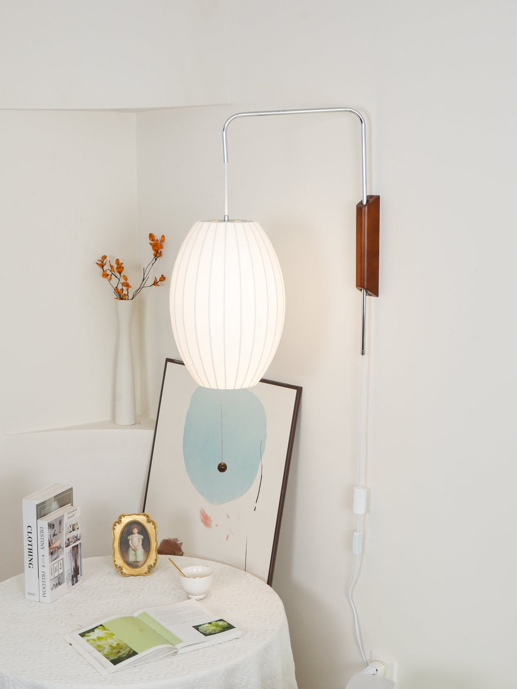 Narrower view of an oval lit Clérachic wall light in a cream niche, above a table with books, a gold frame and a framed print.