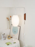 Narrower view of an oval lit Clérachic wall light in a cream niche, above a table with books, a gold frame and a framed print.