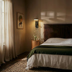 Clérachic black wall reading light lit beside a dark wooden headboard, above a round oak bedside table with white lilies.