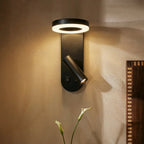 Clérachic black wall reading light with ring panel and spotlight both lit, on a warm wall beside a calla lily stem.
