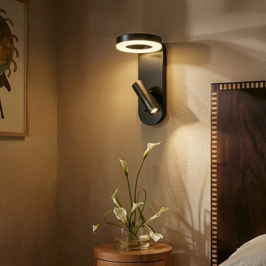 Clérachic black wall reading light lit above a round wooden bedside table of calla lilies, next to a walnut headboard.
