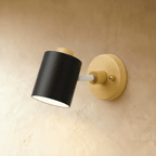Clérachic wall lamp with a matte black cylindrical metal shade and white arm on a light wood backplate, on a limewash wall.