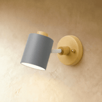 Clérachic wall lamp with a slate grey cylindrical metal shade on a pale wood round backplate, angled on a limewash wall.